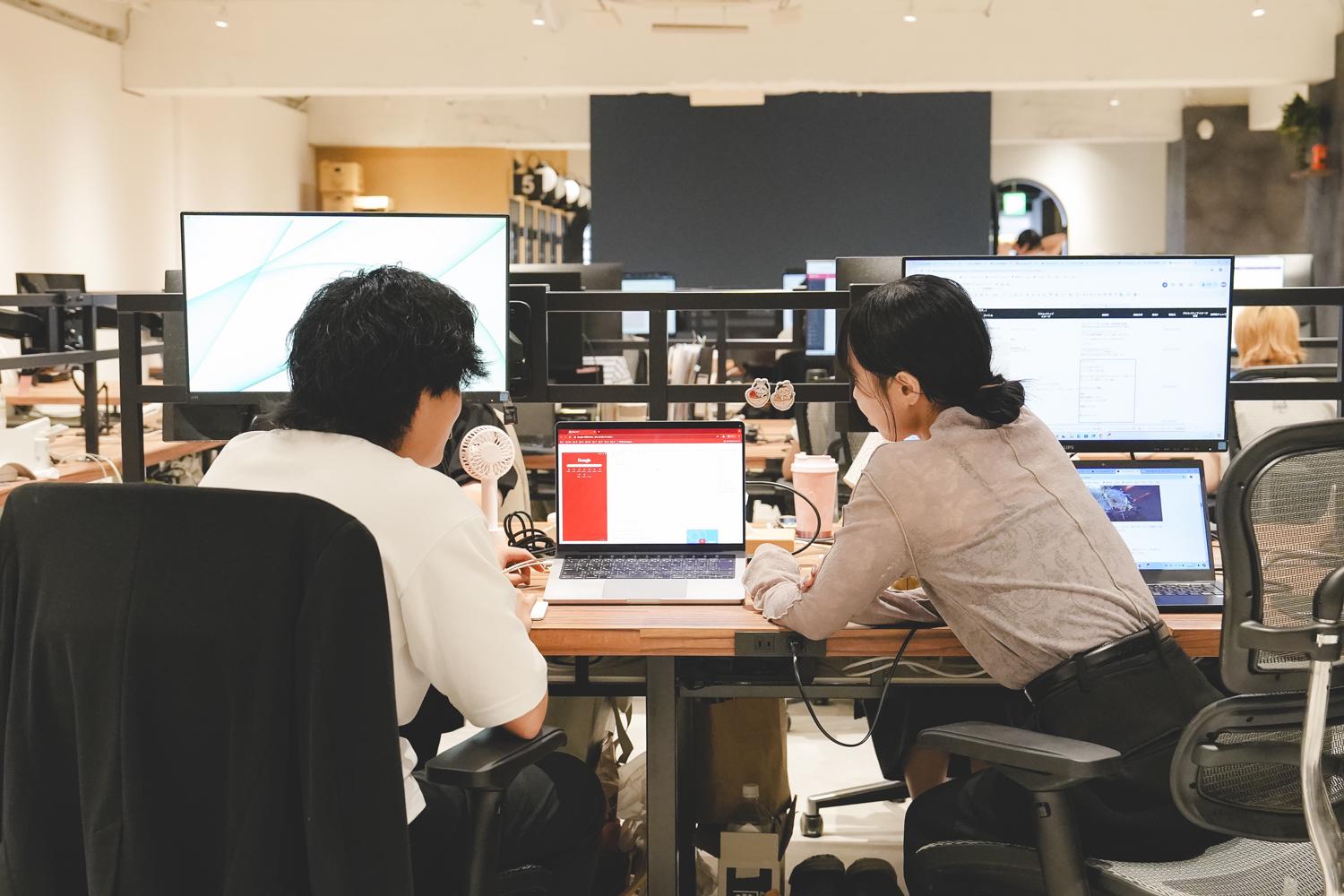 デスクで向き合って仕事する二人。複数のモニターを使って、協力しながら作業している様子が伝わる職場風景。