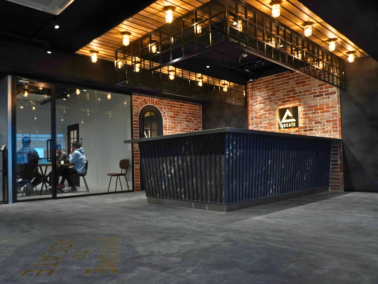 温かな照明とレンガ壁が特徴的なアドケイトのレセプションエリア。黒い波型カウンターと木格子天井が印象的な洗練された空間。