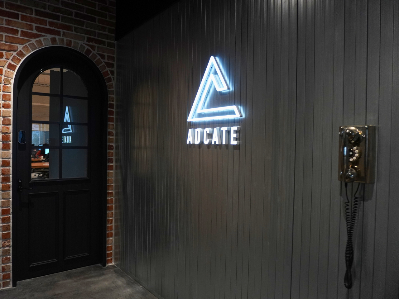 アドケイトのネオンロゴが輝くダークトーンのエントランス。アーチ型ドアとレンガ壁が印象的なモダンなオフィス入口。